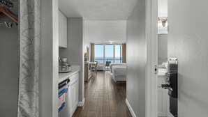 Un pasillo estrecho con una cocina a un lado, un comedor con mesa y sillas, y un dormitorio con cama al otro lado.