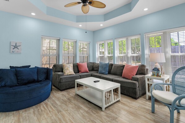A living room with a blue sofa, a gray sectional sofa, and a white coffee table. There are large windows with blinds, a ceiling fan, and a small table with a lamp.
