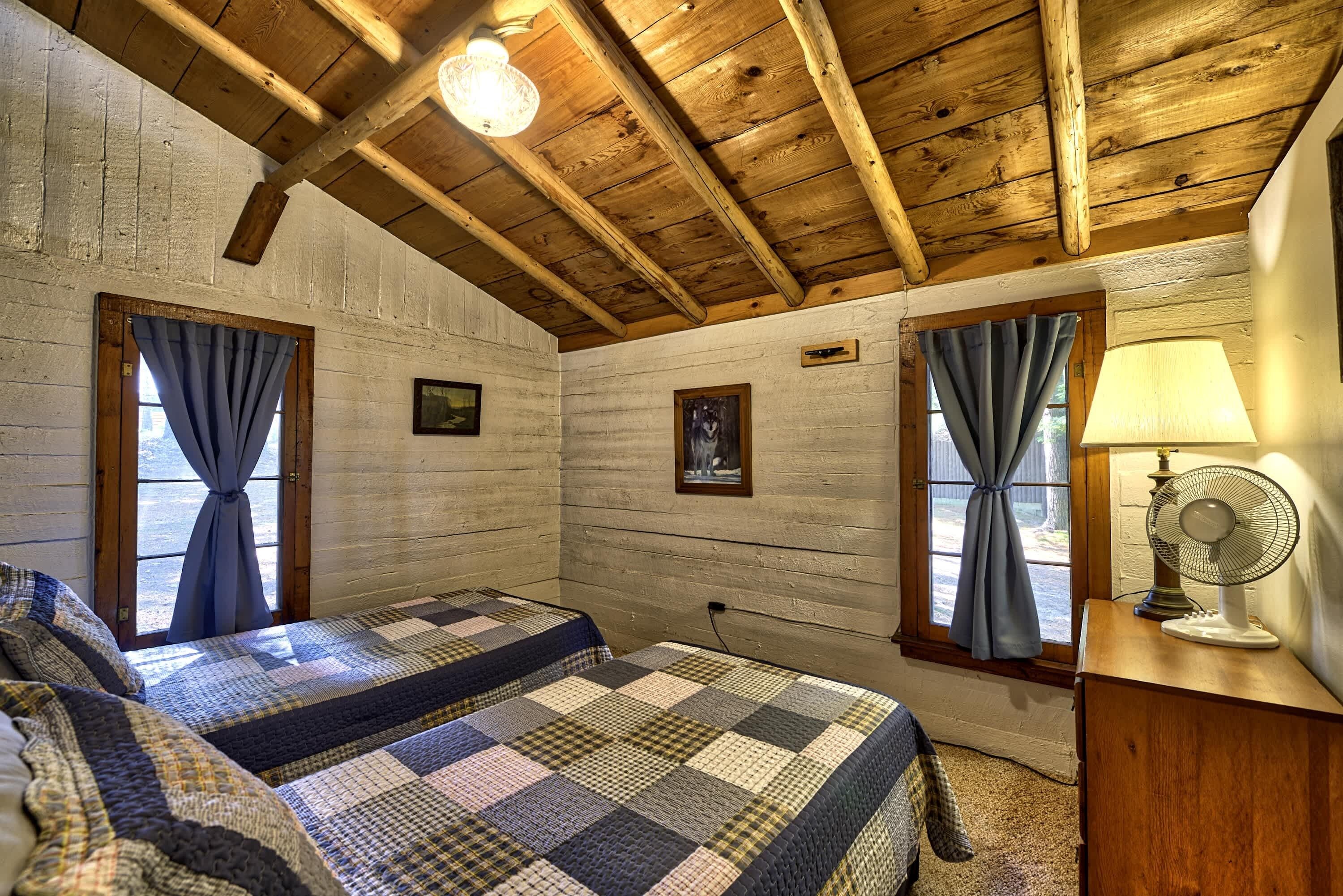 Ein Schlafzimmer mit Holzbalken, zwei Betten mit Decken, einem Ventilator und einem Fenster mit Vorhängen.