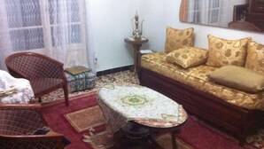 A living room with a sofa, a patterned armchair, a small table with a tablecloth, and a decorative lamp. - Vacation room (El Biar)