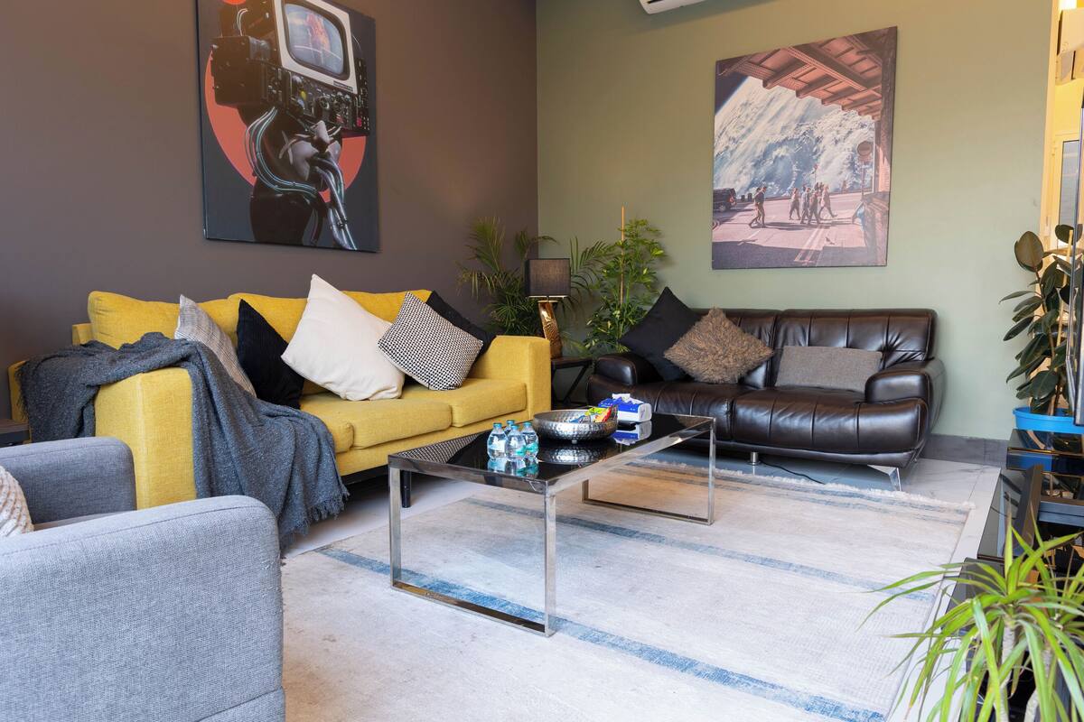 A living room with a yellow sofa, a black leather couch, and a glass coffee table. There are two paintings on the walls and a lamp on a side table.