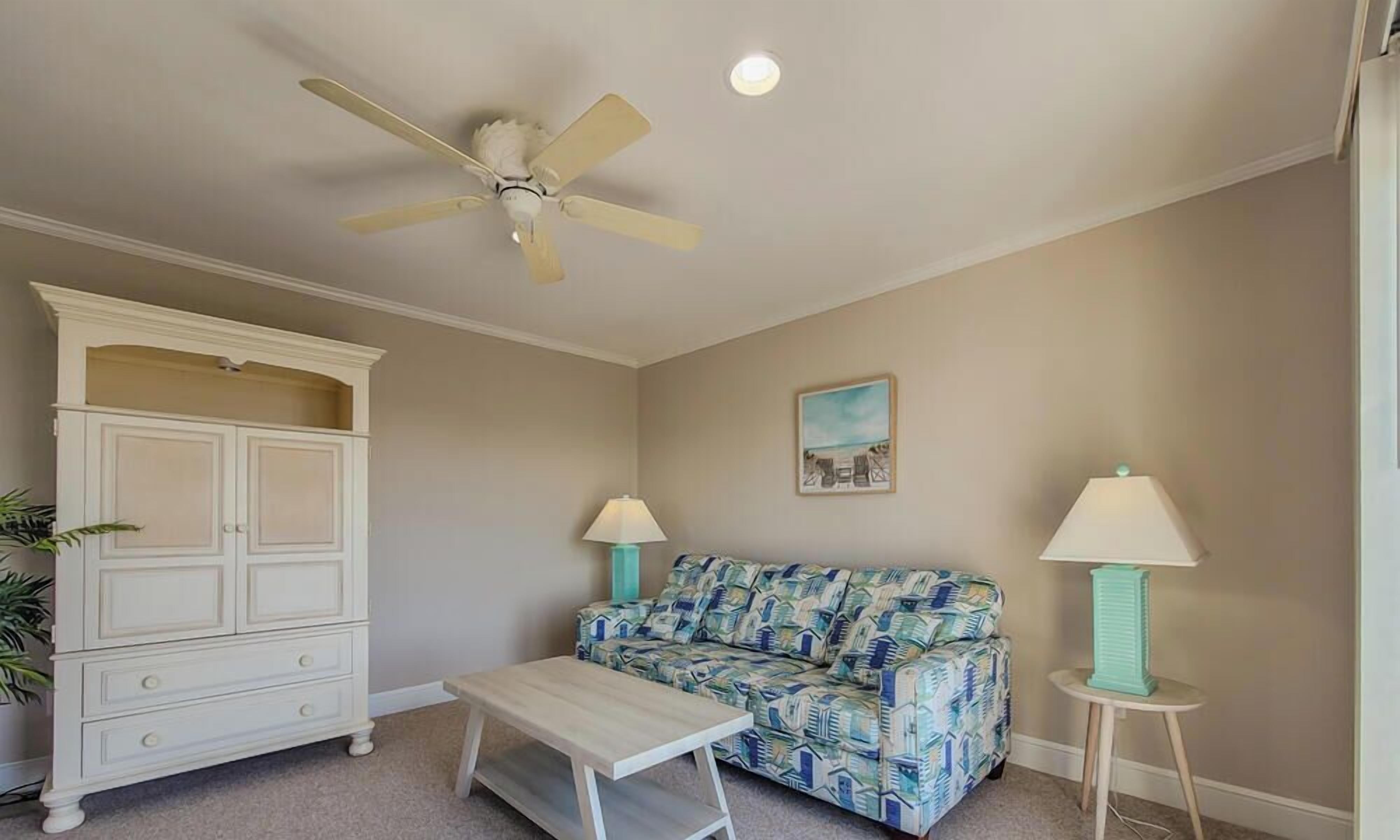 A room with a ceiling fan, a white cabinet, a patterned sofa, a small table, and two lamps.