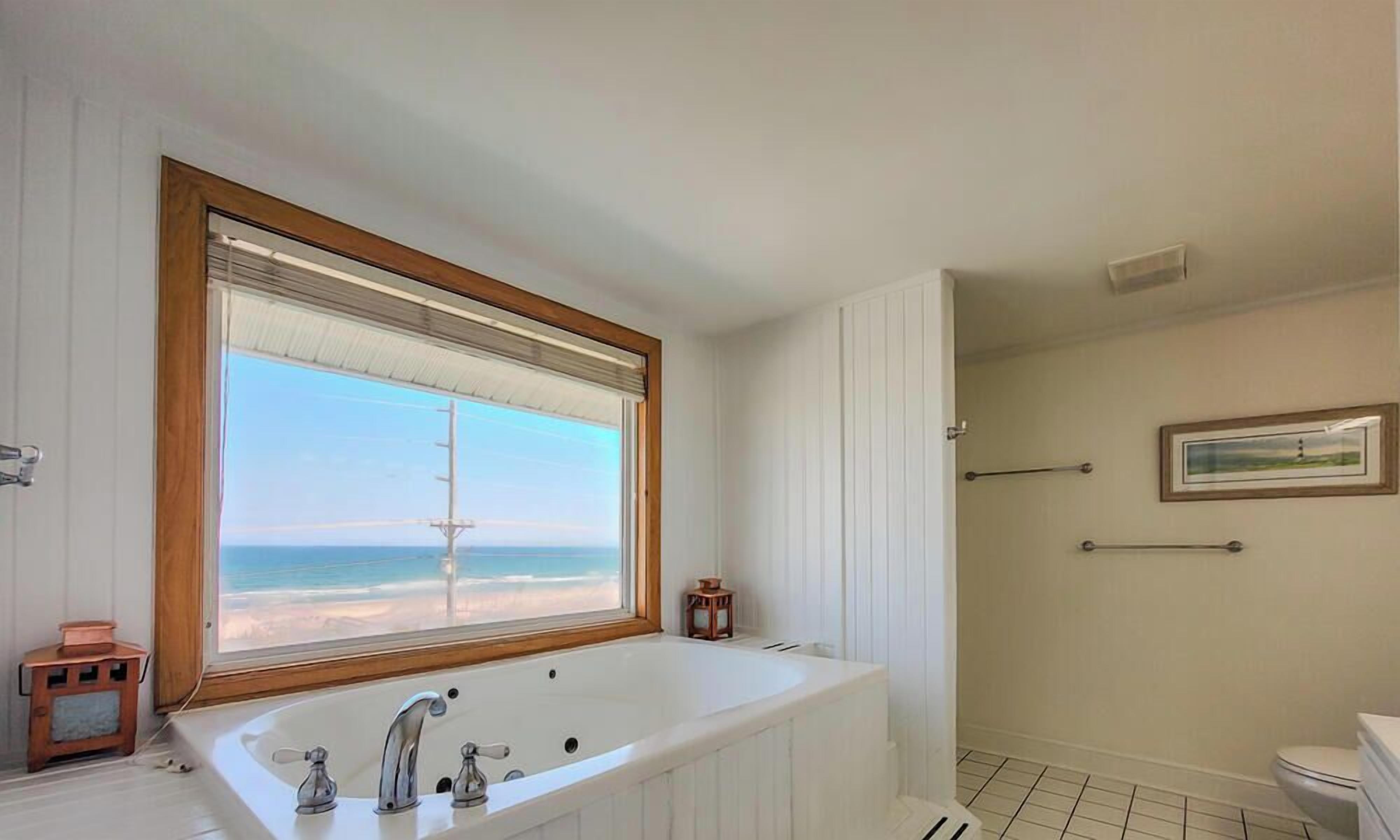 A bathroom with a large bathtub, a window with a view of the beach, a toilet, and a shower area.