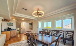A dining area with a wooden table and chairs, a ceiling fan, and a view of the outside through large windows. - Oceanside Manor – High Tide: Beachside Luxury (Surf City)