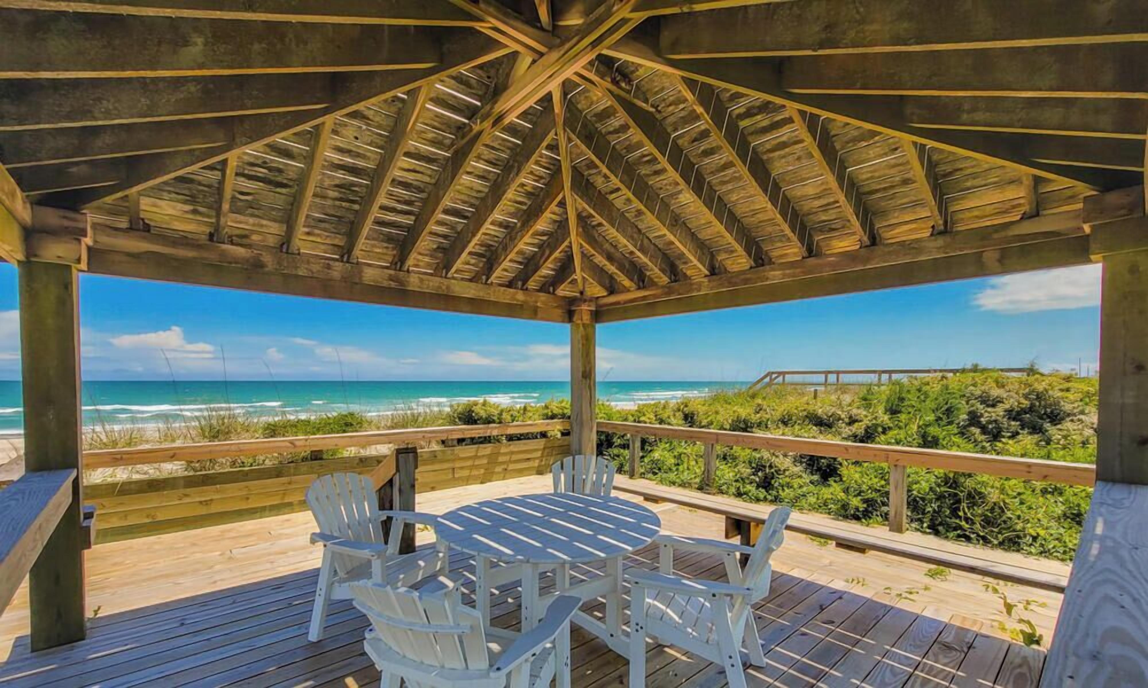 A covered outdoor area with a wooden deck, white chairs, and a table, overlooking a beach and ocean.