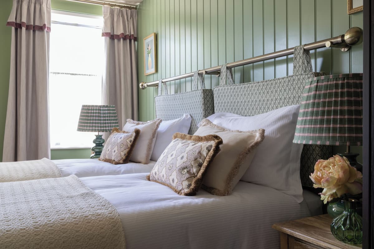 A bedroom with a bed, pillows, a nightstand, a lamp, and a window with curtains.
