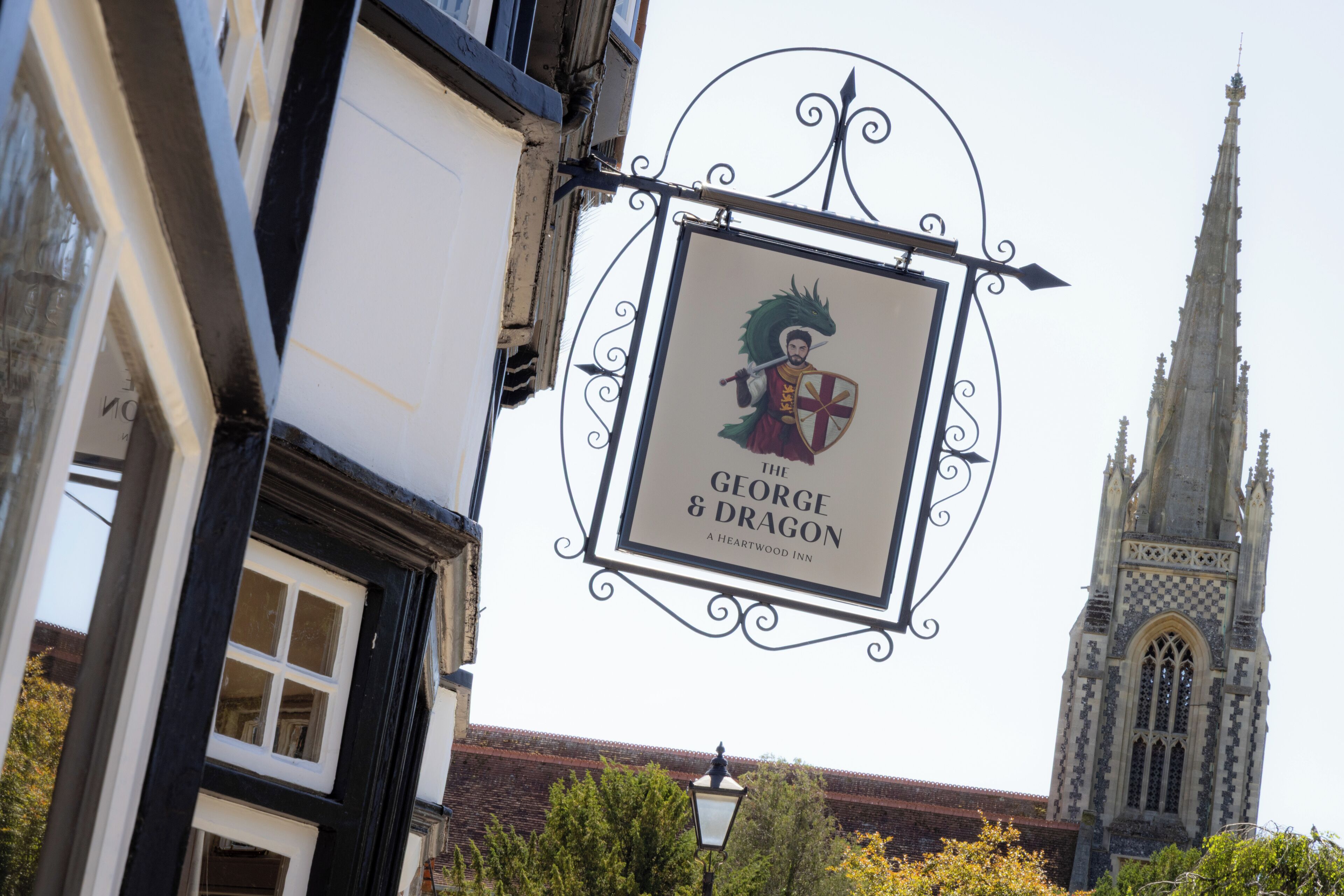 A sign for "The George & Dragon" pub with a knight illustration, set against a backdrop of a church tower and a building with a black and white facade.