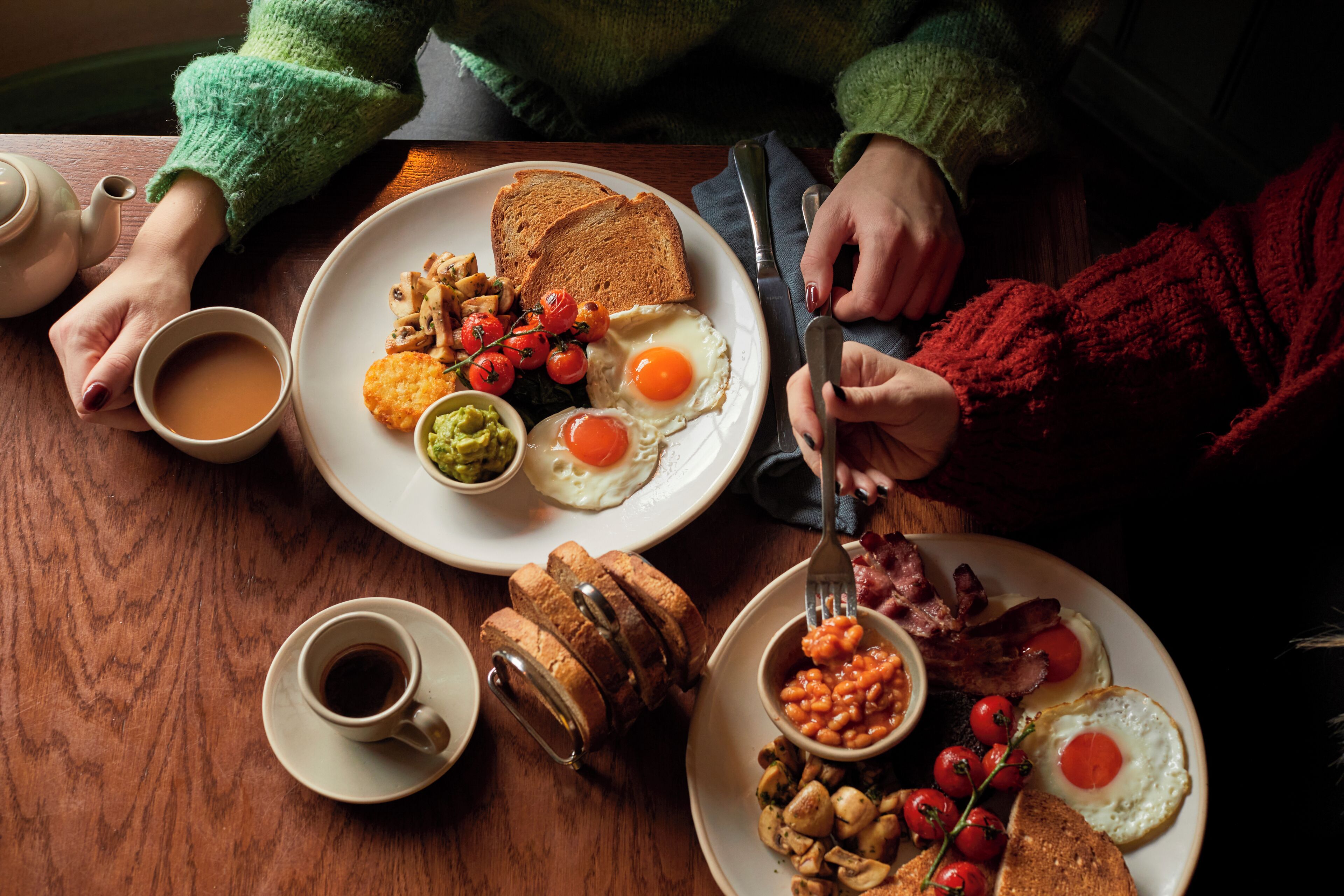 A wooden table with breakfast plates containing eggs, toast, beans, and tomatoes.