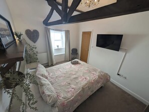 A bedroom with a bed, a TV mounted on the wall, a window with curtains, and a radiator. - Hocking Cottage (Torquay)