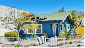 Una casa de dos pisos con fachada azul y techo verde, rodeada por una valla de piquetes blancos y diversas plantas.
