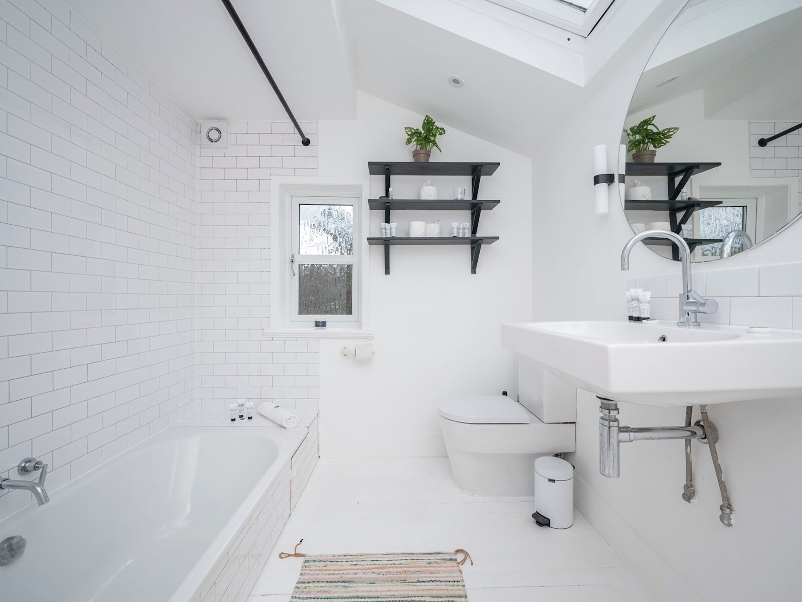 A modern bathroom with a bathtub, toilet, and sink. There are two shelves with toiletries and a window above the toilet.