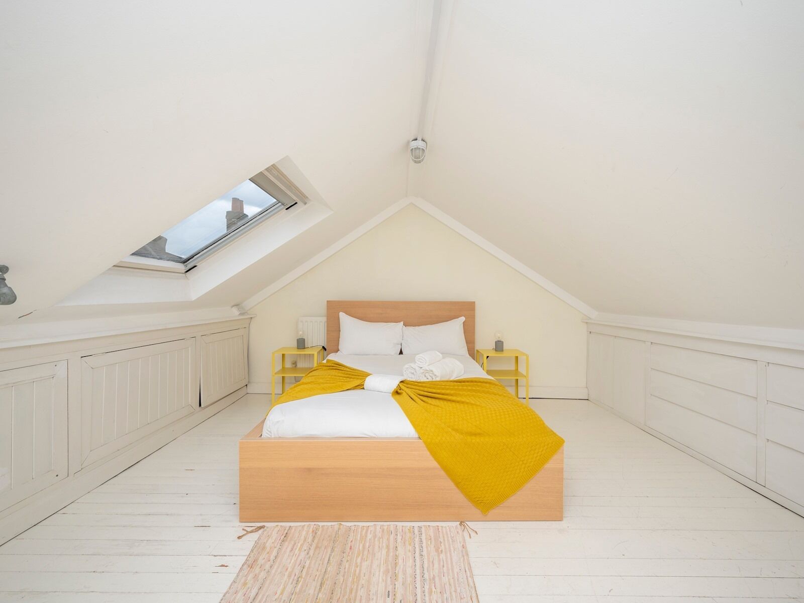 Un dormitorio con techo inclinado, tragaluces, cama con ropa de cama blanca y un plaid amarillo, cabecero de madera y paredes blancas.