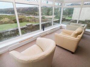 A room with a large window overlooking a river and greenery, a beige sofa, and a small table. - 1 Atlantic View (Newquay)