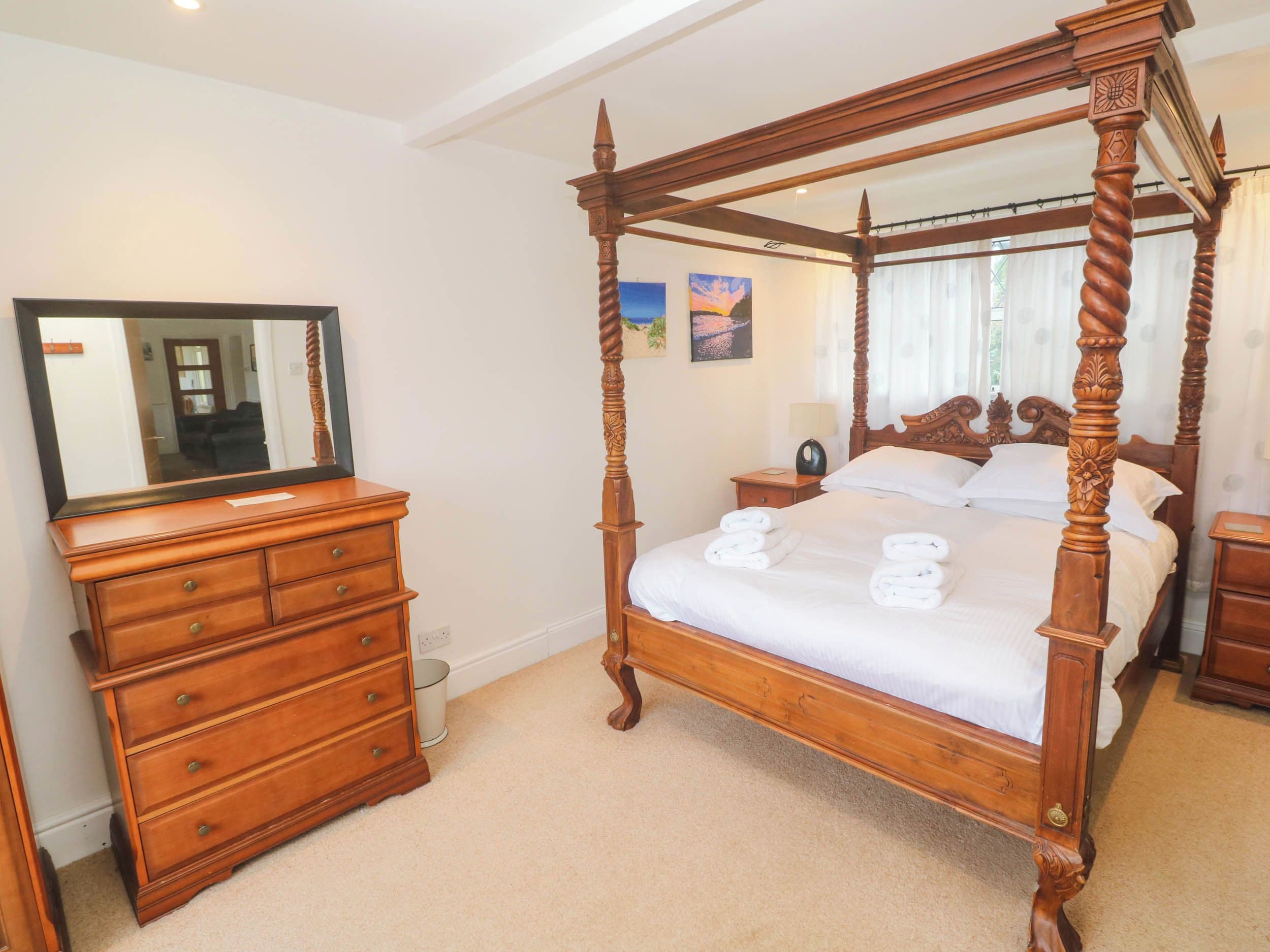 Un lit à baldaquin avec une structure en bois et du linge de lit blanc, une commode en bois avec des tiroirs, une table de chevet avec une lampe et un miroir posé sur la commode.