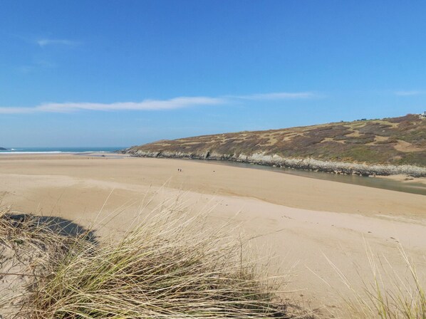 A wide sandy beach with gentle waves, a grassy dune, and a clear blue sky.