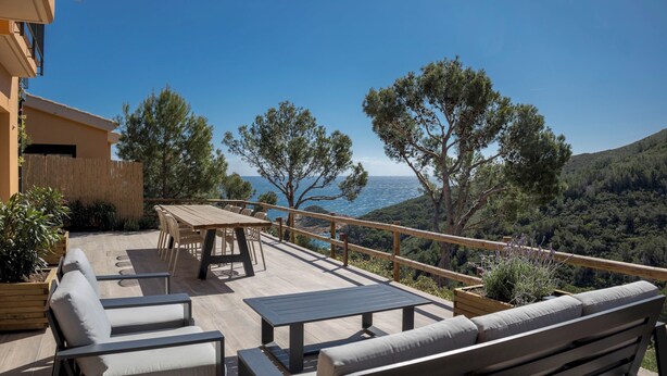 An outdoor deck with seating and a table, overlooking a mountainous landscape and the sea.