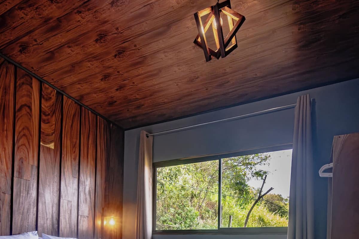 Une chambre avec un plafond en bois, un lit recouvert d’une courtepointe, une table de chevet, une fenêtre donnant sur la verdure et un luminaire suspendu.