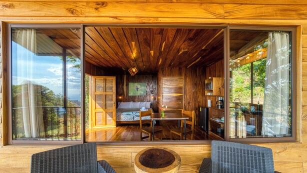 Una cabaña de madera con comedor, cocina y un amplio ventanal que ofrece vistas.