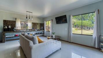 Ein modernes Wohnzimmer mit einer Couch, einem Essbereich mit Küchenzeile, einem an der Wand montierten Fernseher und großen Fenstern mit Vorhängen.