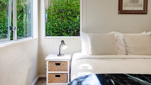 Une chambre à coucher avec un lit, une table de chevet avec une lampe, deux paniers en osier, une fenêtre donnant sur la verdure et un tableau encadré au mur.