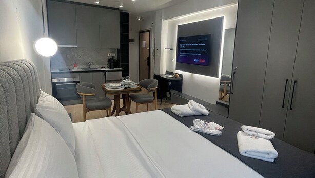 Ein modernes Hotelzimmer mit einem ordentlich bezogenen Bett, einem kleinen Schreibtisch mit Stuhl, einem an der Wand befestigten Flachbildfernseher und einer Küchenzeile im Hintergrund.