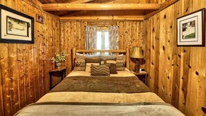 Ein Schlafzimmer in einer Holzhütte mit einem Bett, Kissen, einem Nachttisch, einer Lampe und gerahmten Bildern an den Wänden.