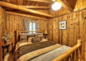 Un dormitorio de cabaña de madera con cama, almohadas, una mesita de noche, una lámpara, una ventana con cortinas y un cuadro en la pared.