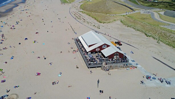 An aerial view of a beach with numerous umbrellas and people. A large building with a red roof and white walls is located near the center. - Holiday Home Near Petten Beach With Pool (Petten)