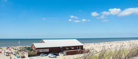 A beachside building with a flat roof, adjacent to a sandy beach with people and umbrellas.