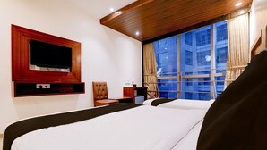 Une chambre d’hôtel avec deux lits, un téléviseur fixé sur un panneau en bois, une fenêtre avec des rideaux et une petite table avec une chaise.