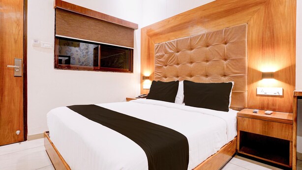 Una cama bien tendida con cabecera de madera, dos mesitas de noche con lámparas y una ventana con persianas.