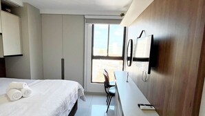 A modern hotel room with a bed, a desk, a chair, a TV, and a window with a city view. - Cayenne By Day by Day (Joao Pessoa)