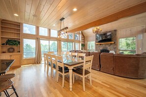 Un salón comedor amplio con piso de madera, una mesa de vidrio rodeada de sillas, un sofá seccional marrón, una chimenea de piedra y un televisor de pantalla plana montado en la pared.