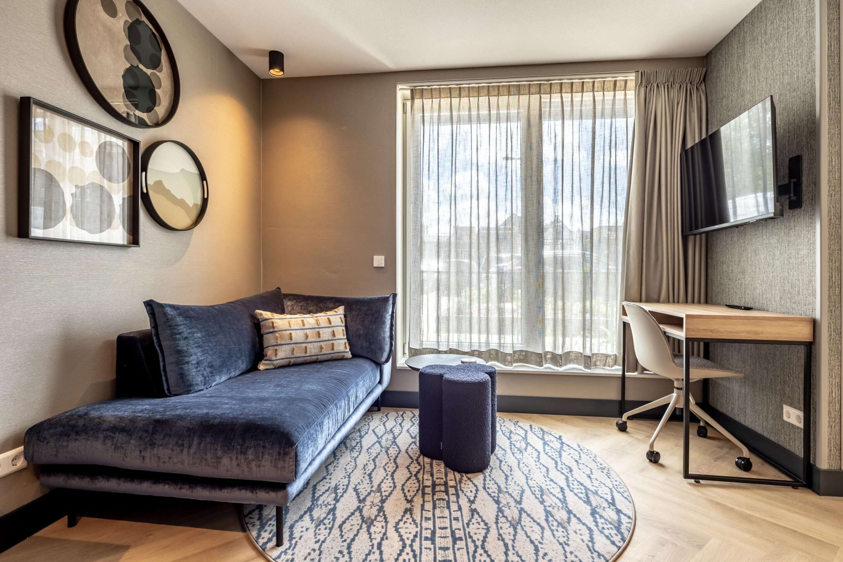 Una sala de estar moderna con un sofá azul oscuro, una alfombra redonda, un pequeño otomaní y un escritorio con una silla.