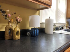 A kitchen counter with two vases of dried flowers, a blue vase, a white paper towel holder, and a window with blinds.