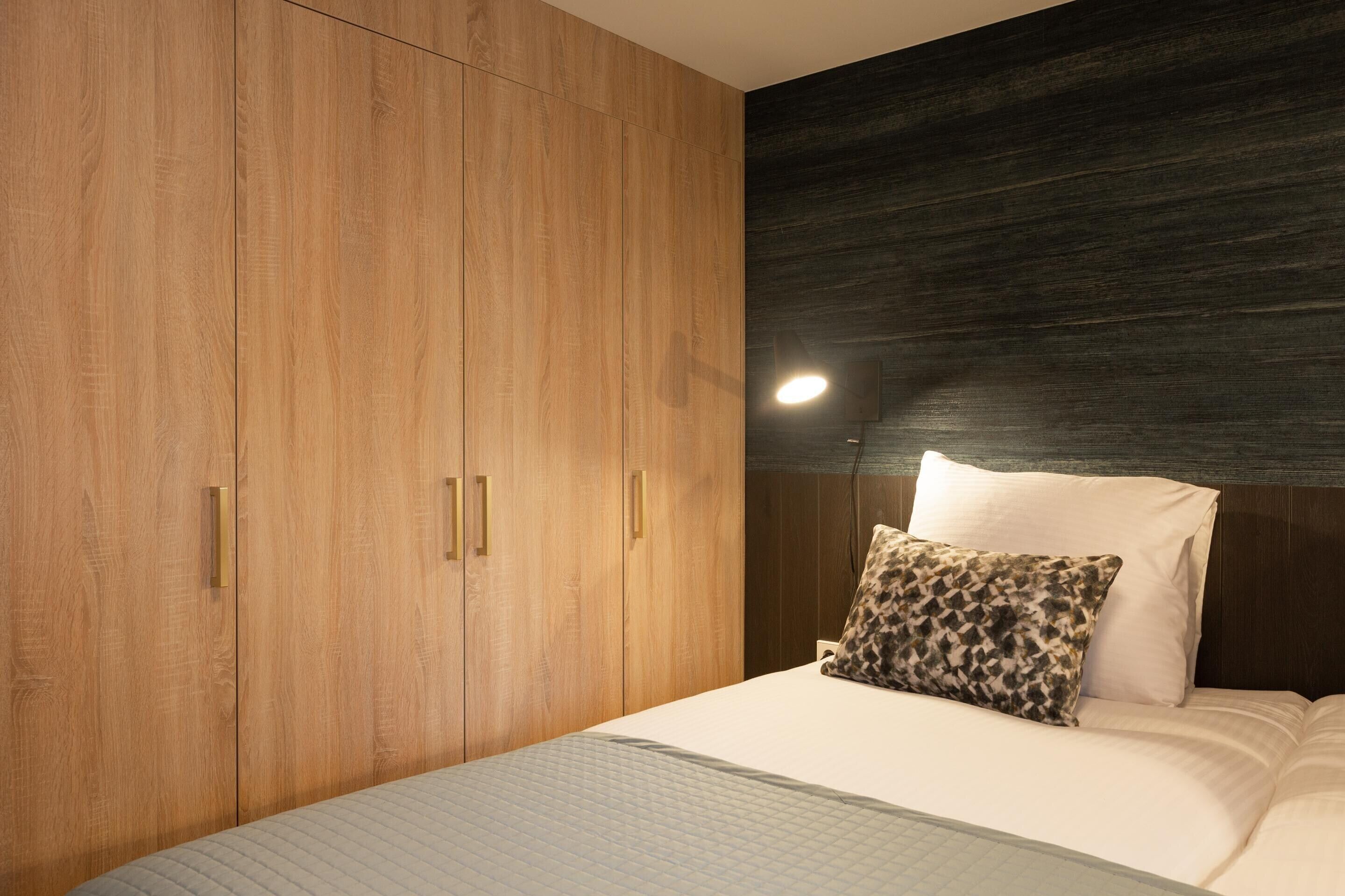 A bedroom with a large wooden wardrobe, a bed with white bedding, and a dark textured wall.