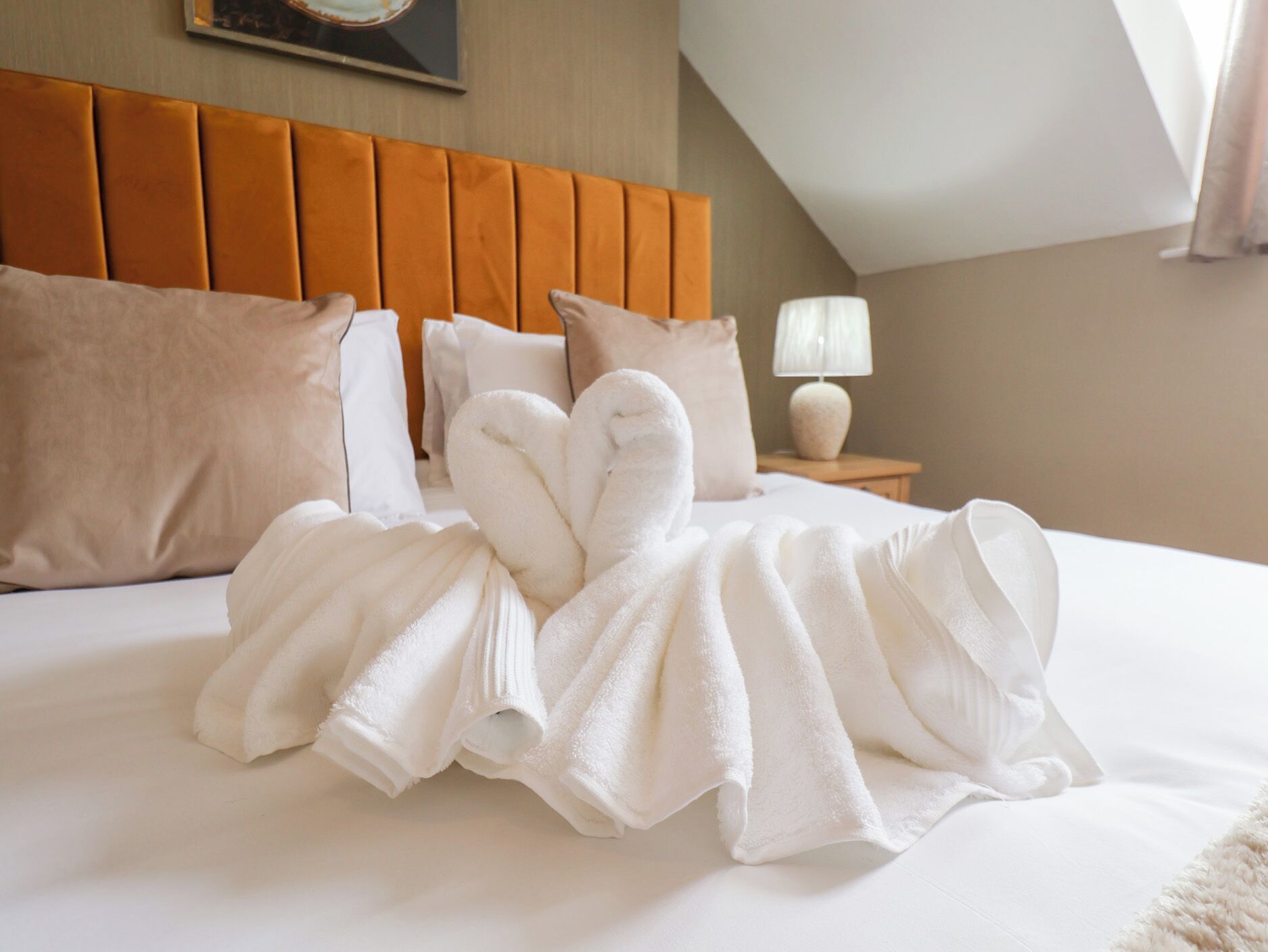 A neatly made bed with white towels arranged in a heart shape, a wooden headboard, and a bedside lamp.