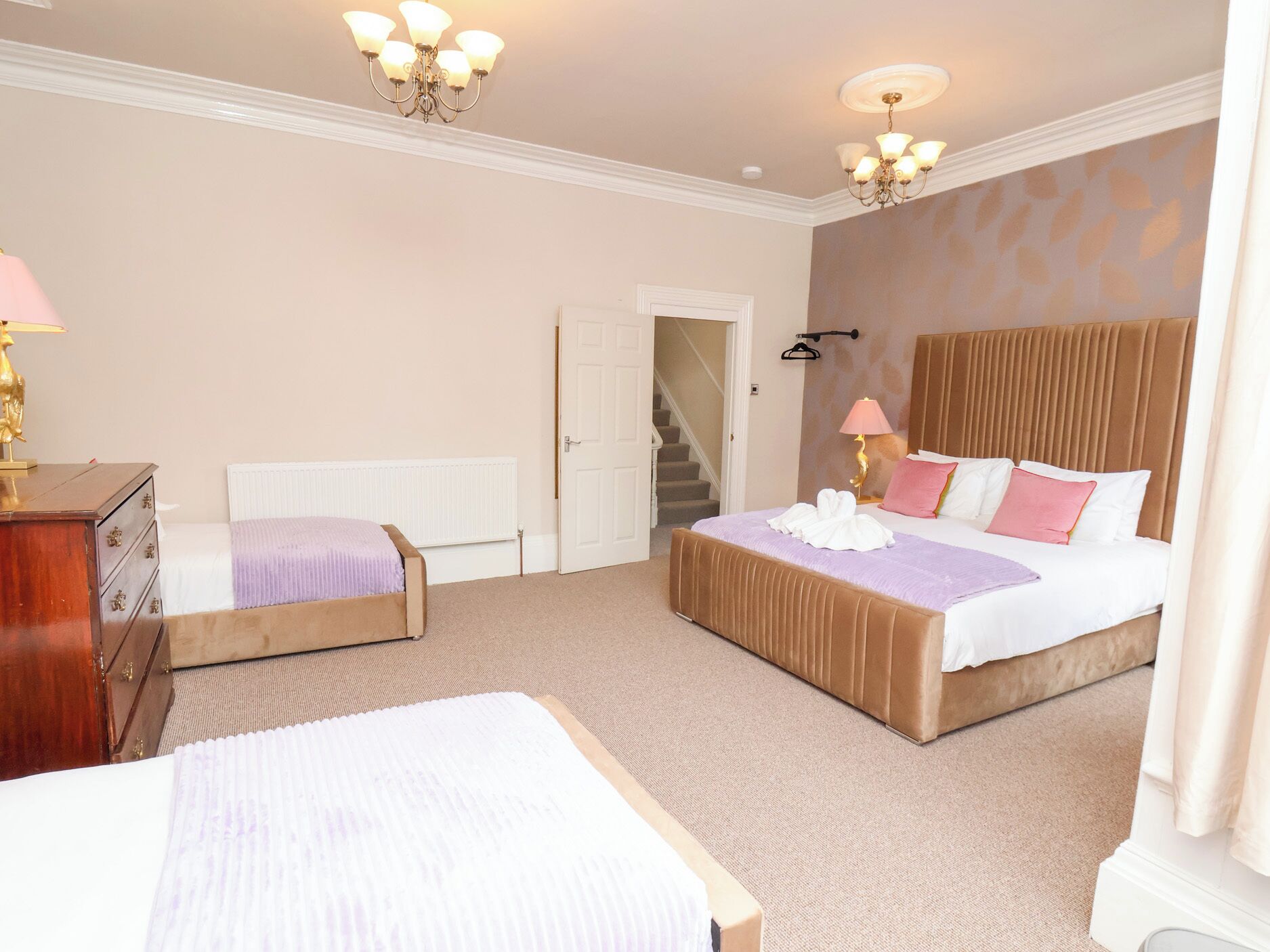 A bedroom with two beds, a dresser, a radiator, a chandelier, and a staircase.