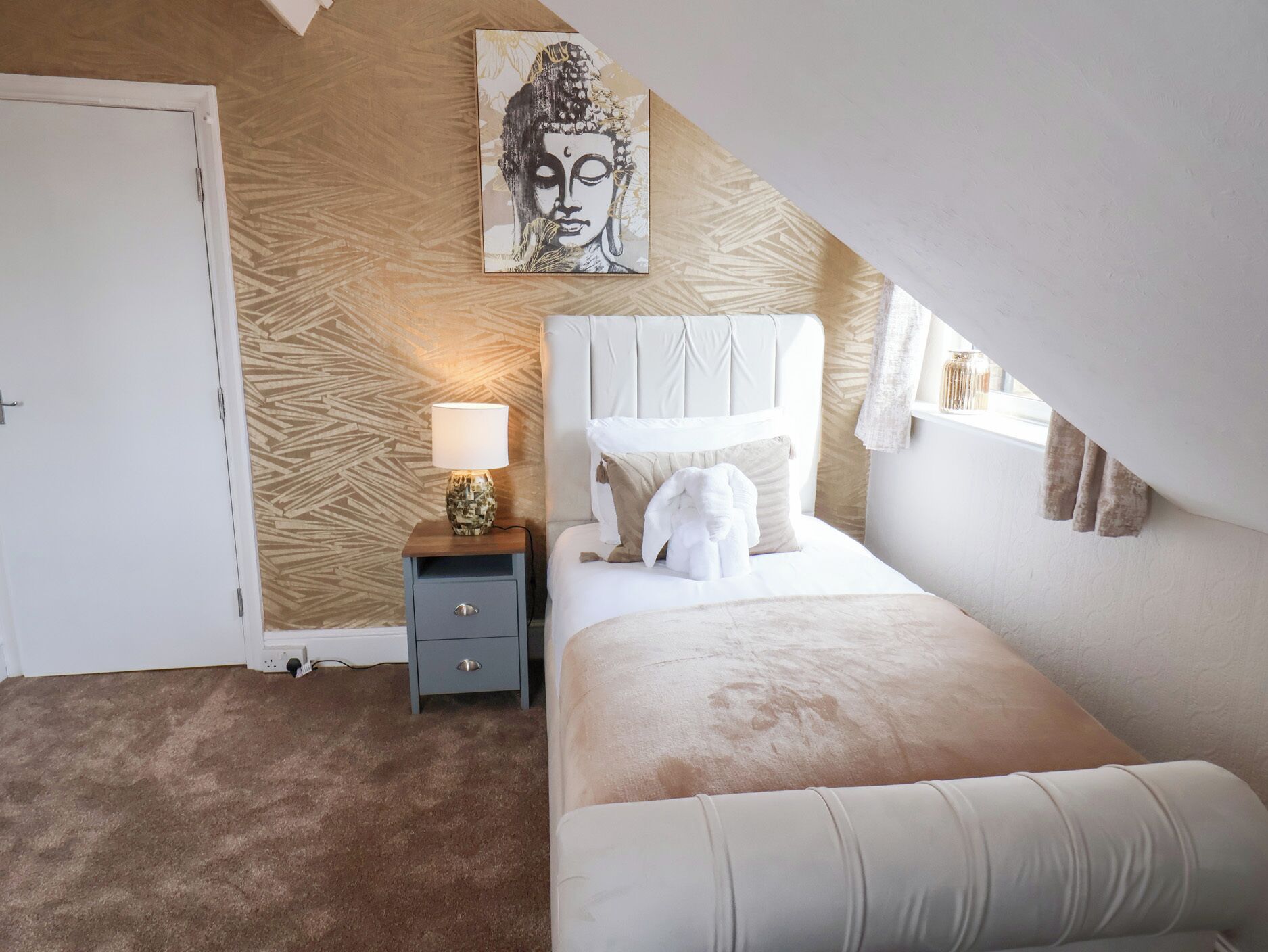 Un dormitorio con cama, mesita de noche, lámpara, una obra de arte en la pared y ventana con cortinas.