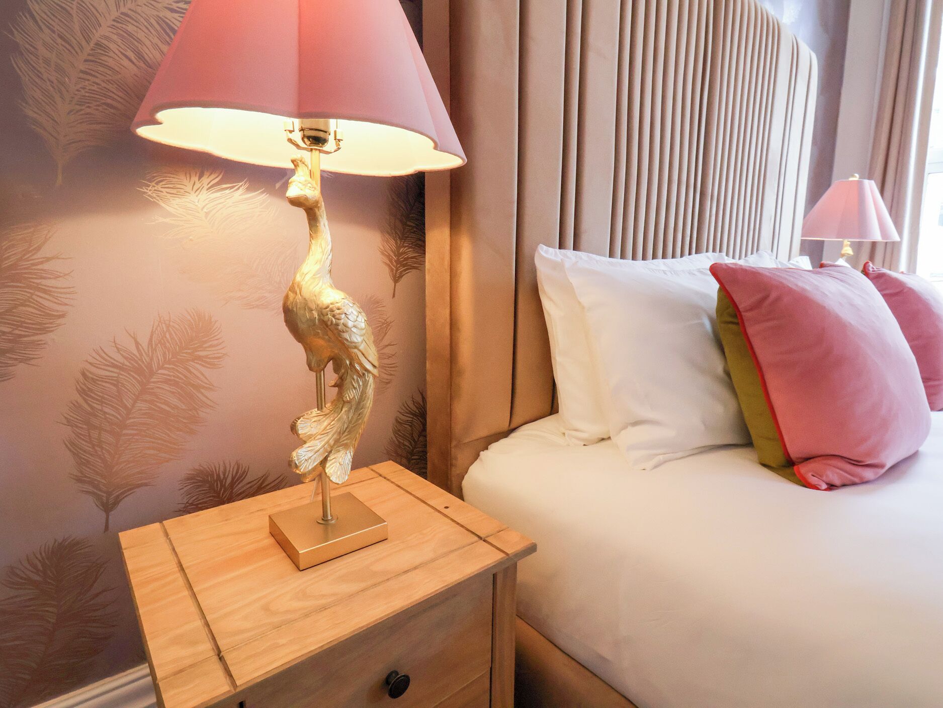 Un lit bien fait, avec des oreillers blancs et roses, une table de chevet en bois surmontée d’une lampe, et un mur orné d’un motif de feuilles.