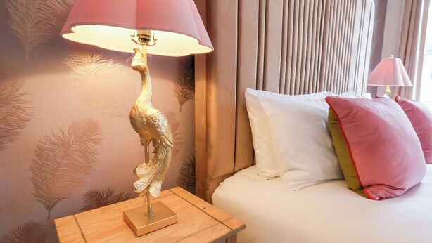 Un lit bien fait, avec des oreillers blancs et roses, une table de chevet en bois surmontée d’une lampe, et un mur orné d’un motif de feuilles.