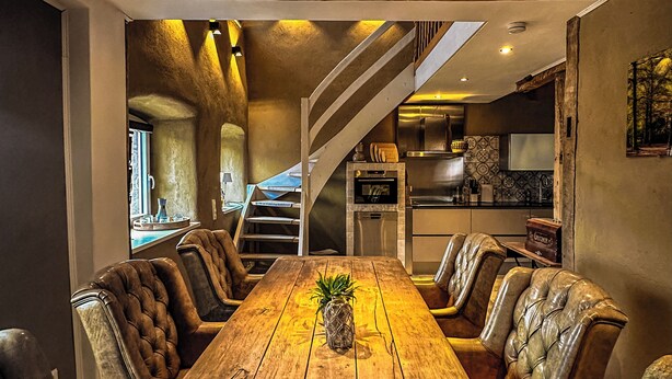 Ein rustikales Esszimmer mit Holztisch und Ledersesseln, einer Treppe zu einer oberen Ebene sowie einer Küche im Hintergrund.