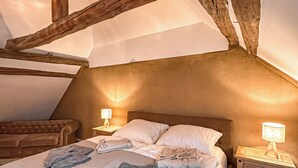 Ein gemütliches Schlafzimmer im Dachgeschoss mit schrägen Wänden, freiliegenden Holzbalken, einem großen Bett mit weißer Bettwäsche, zwei Nachttischen mit Lampen und einem braunen Ledersofa.