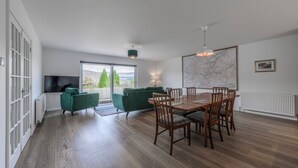 A spacious living area with a dining table, green sofas, and a large window offering a view of trees and mountains.