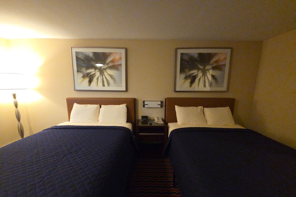 Ein Hotelzimmer mit zwei Betten, jeweils mit Kopfteil, Nachttisch, Lampe und zwei gerahmten Bildern an der Wand.