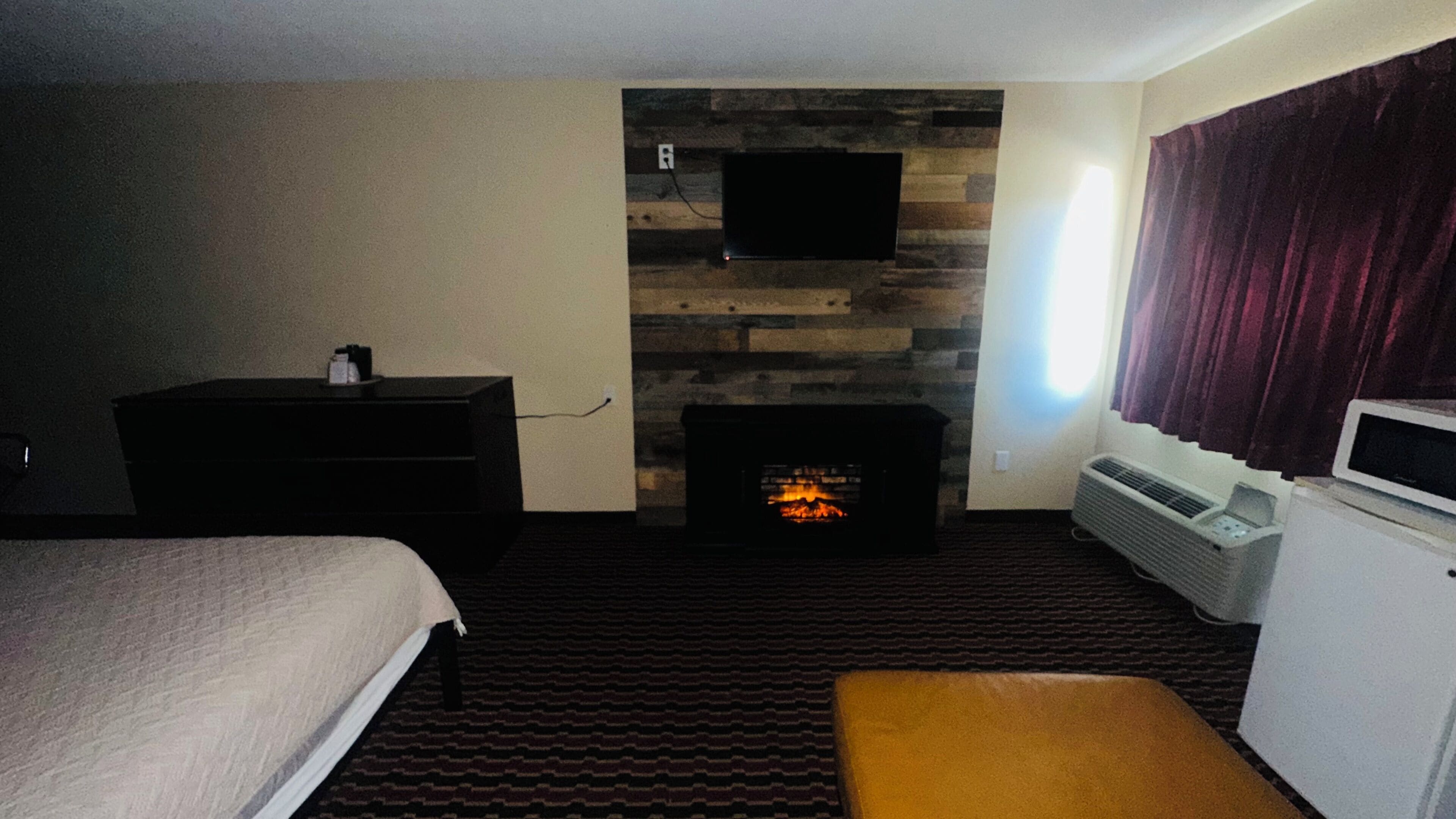 Une chambre d’hôtel comprenant un lit, une commode, un téléviseur à écran plat fixé à un mur en bois, une cheminée et une fenêtre avec des rideaux.