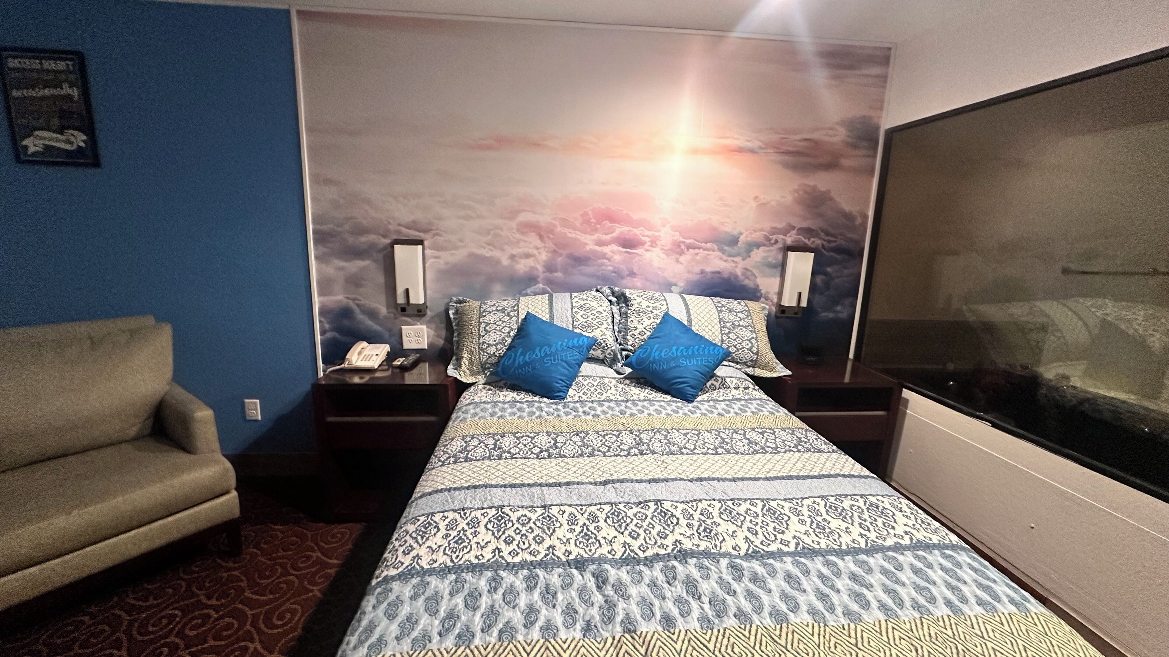 Hotellihuone, jossa on kuviollinen päiväpeite, kaksi sinistä tyynyä, yöpöytä, jossa on puhelin ja lamppu, tuoli ja suuri ikkuna.