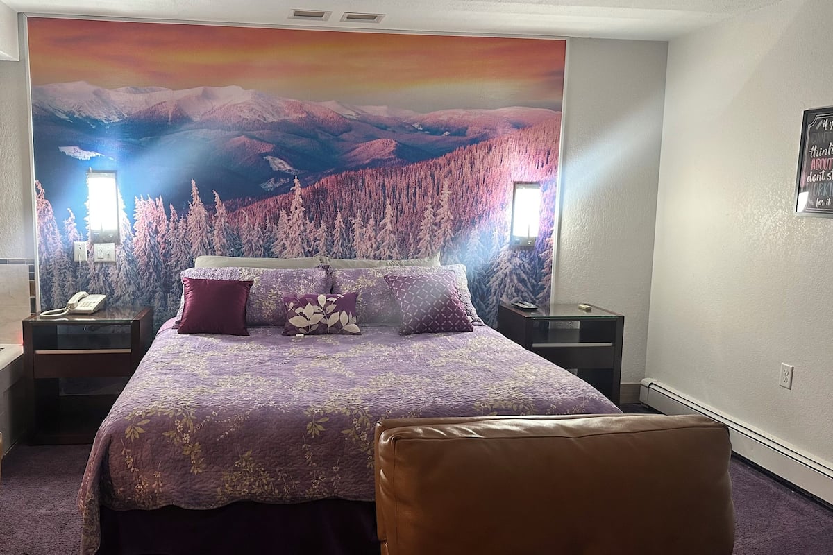 Ein Schlafzimmer mit Wandgemälde von einer Berglandschaft, einem Bett mit violetter Bettwäsche, einem Nachttisch, einem Telefontisch und einem Badezimmer mit einer Badewanne.
