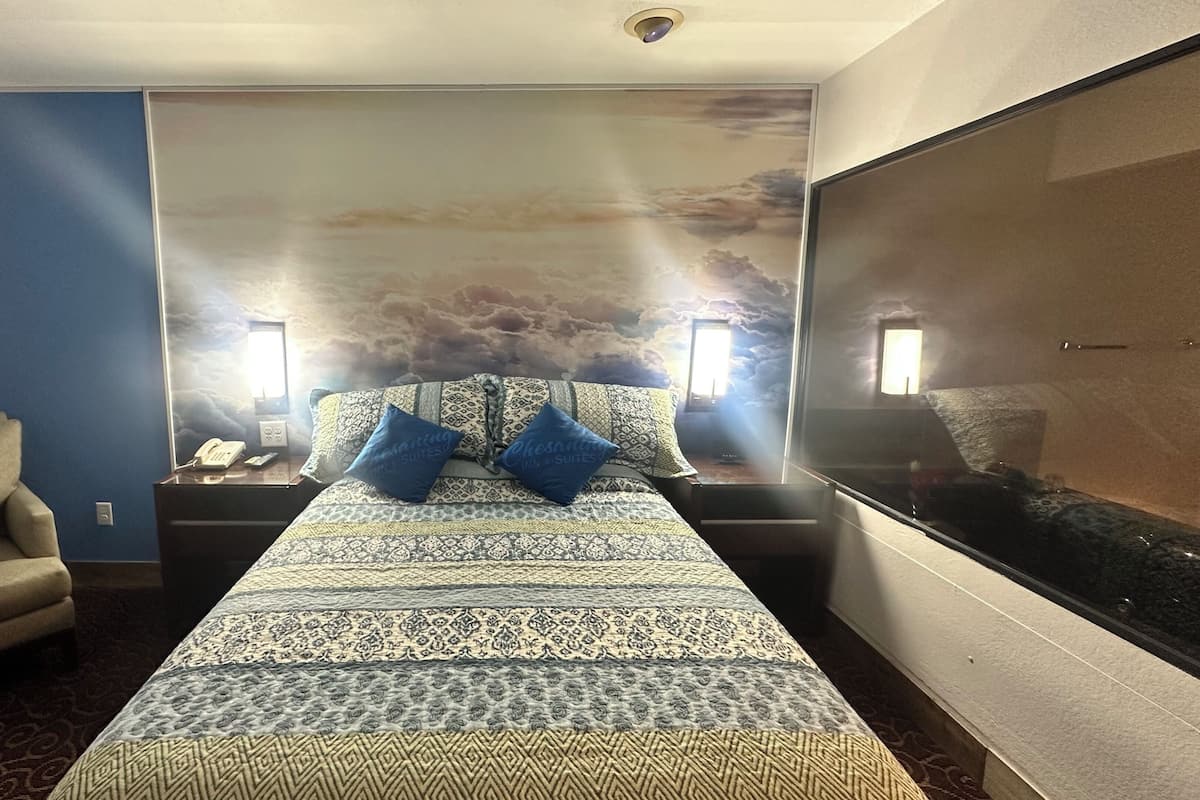 Ein Hotelzimmer mit Bett, einem Sessel, einem Nachttisch mit Telefon und einer Wand mit Wandgemälde von Wolken.