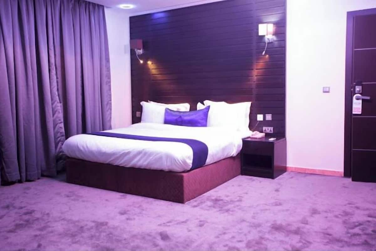 A hotel room with a large bed, a nightstand, a dark wooden headboard, and purple curtains.